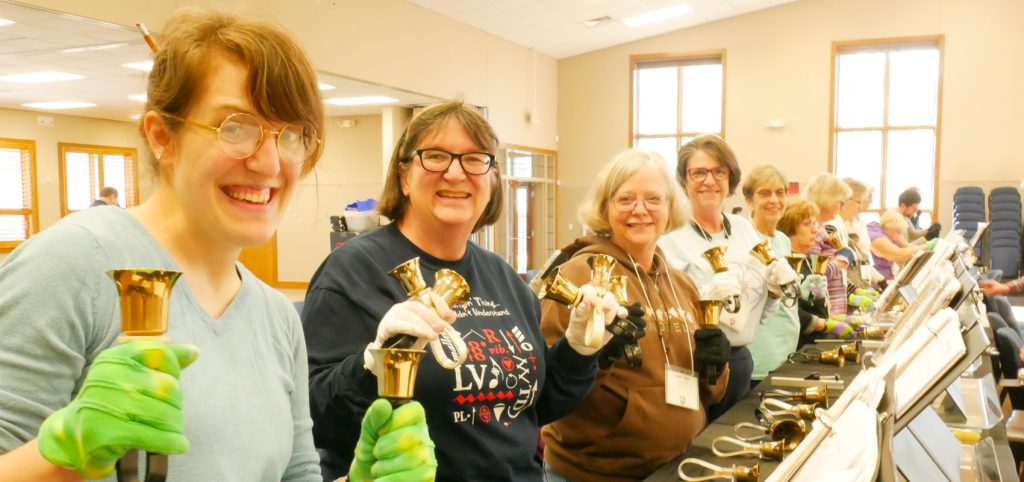 Handbell ringers in green gloves smiling over their bells at a long rehearsal table