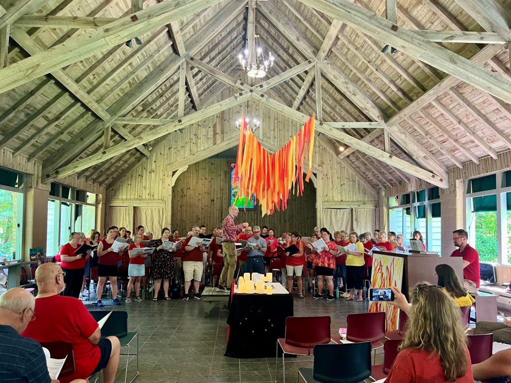 A choir in red shirts sings in a timber-roofed hall at Lutheridge during Music Week, flame-colored streamers hanging above