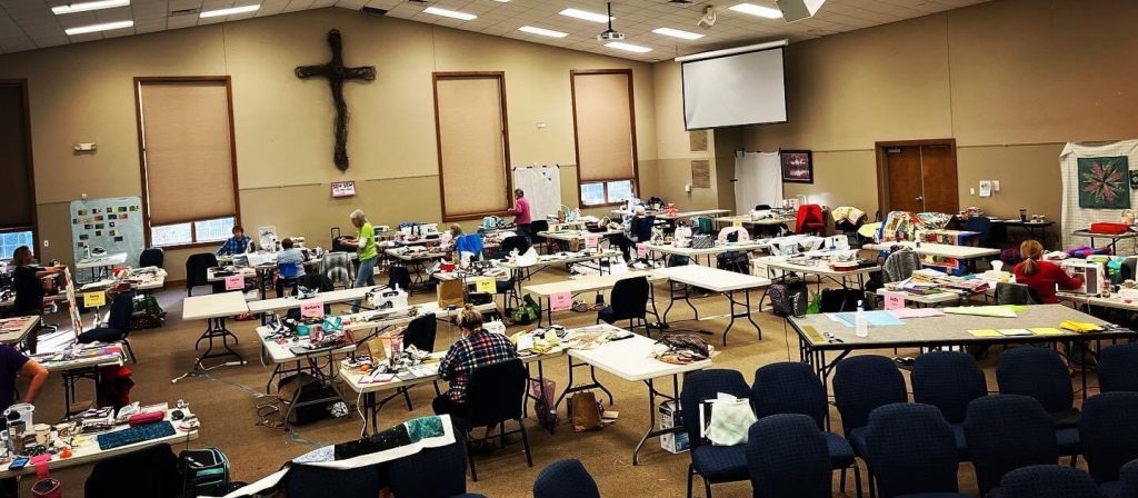Quilters at rows of sewing tables in a large meeting hall with a wooden cross on the wall