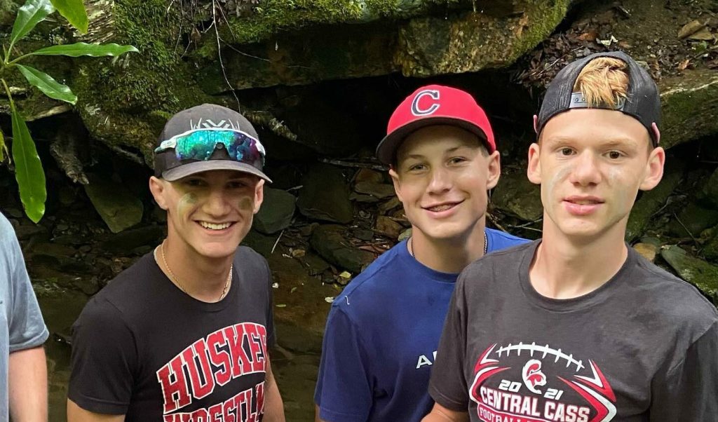 Three teenage campers in caps smile beside a mossy rock ledge on a Lutherock Blue Ridge Expeditions trip