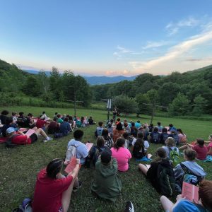 Campers sit on a grassy hillside at Lutherock for outdoor worship, facing a leader who stands among three wooden crosses