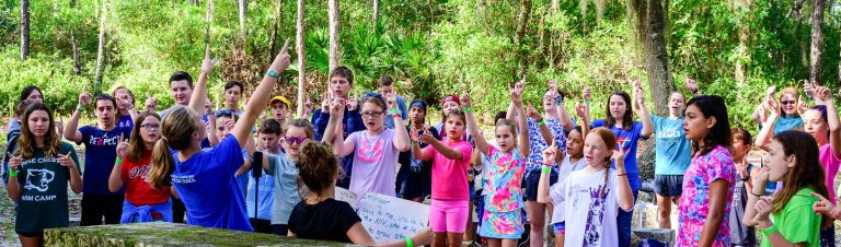 Luther Springs campers sing with hand motions outdoors under the pines