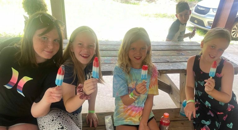 Four smiling girls hold up red, white and blue ice pops at a picnic table at Luther Springs