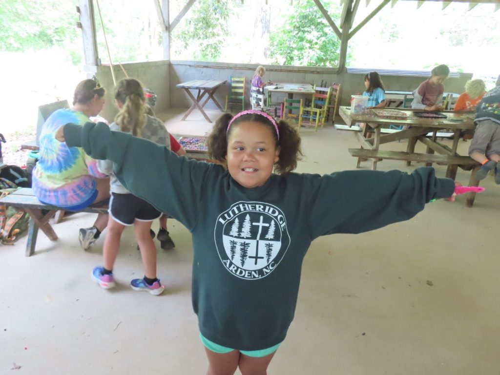 A Lutheridge camper in a Lutheridge sweatshirt flinging her arms wide under the pavilion
