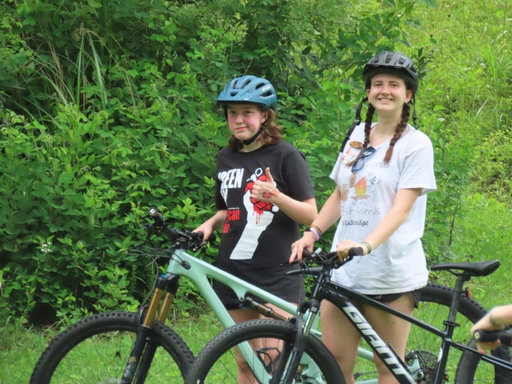 Two teen campers in bike helmets with their mountain bikes on a wooded trail