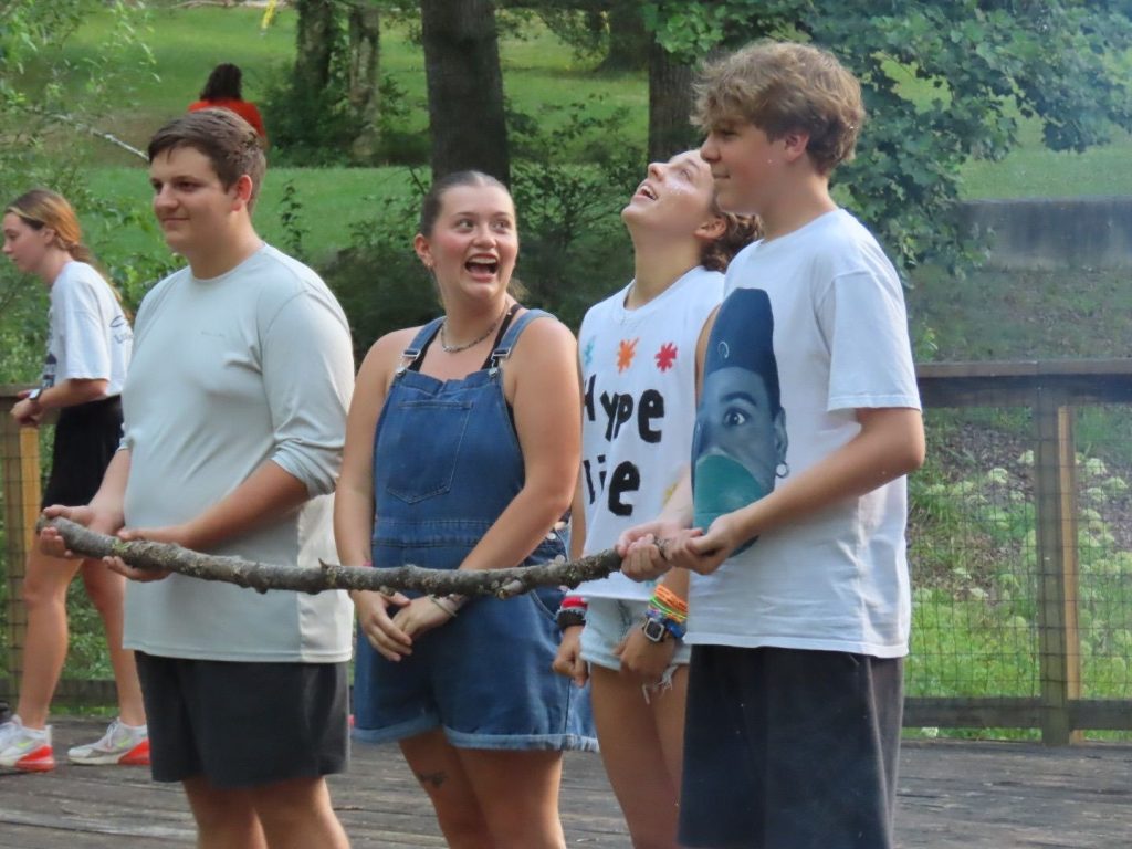 Teen campers at Lutheridge laughing together on a wooden deck, holding a long stick between them