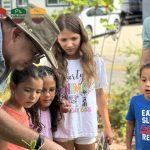 A leader in a bush hat shows a group of young children something in a garden bed