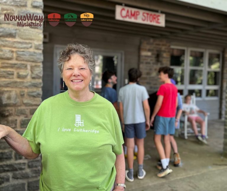 A smiling woman in an I love Lutheridge T-shirt outside the Lutheridge Camp Store, campers on the porch behind her