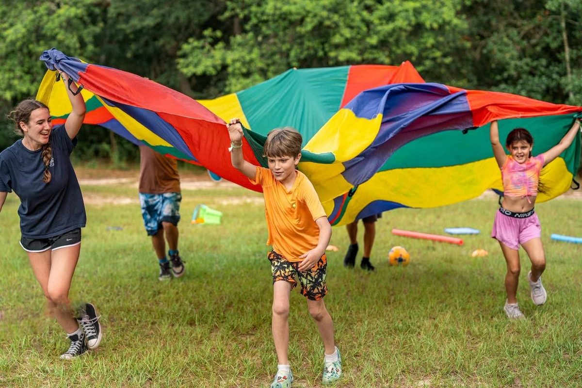 Campers and a counselor running under a rainbow parachute on the field at Luther Springs
