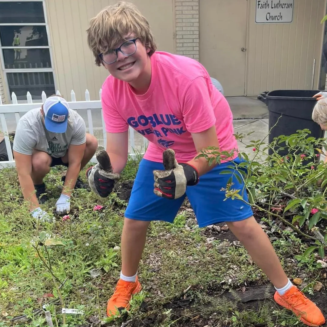 A teen in work gloves giving two thumbs up while clearing a garden bed outside Faith Lutheran Church, an adult working beside him