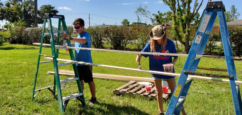 Two volunteers painting trim boards between ladders in a yard on a Luther Springs disaster response trip