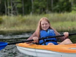 A smiling camper paddles an orange kayak on the lake at Luther Springs
