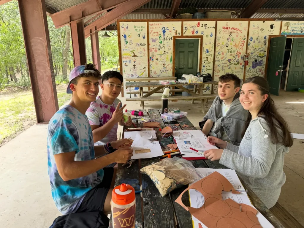 Four teen campers smiling at a picnic table under the Luther Springs pavilion, working on a craft