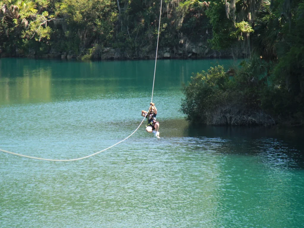 A camper riding a zip line over clear blue-green spring water lined with trees