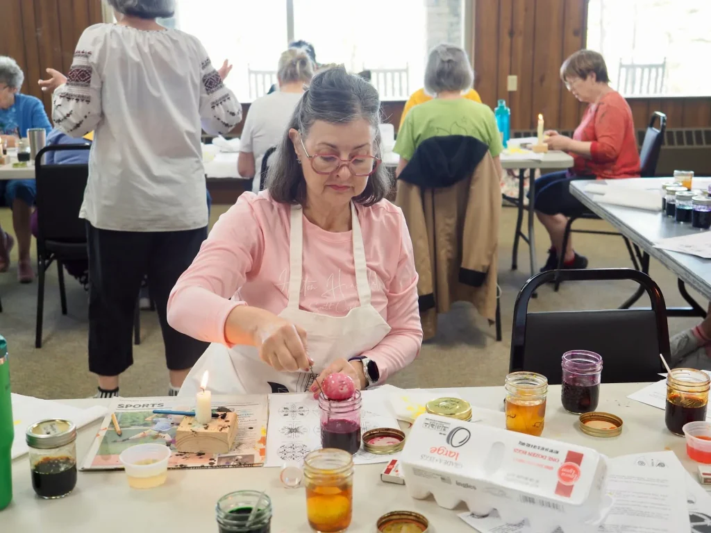 A retreat guest in an apron decorates a dyed egg with wax beside a lit candle, jars of dye around her
