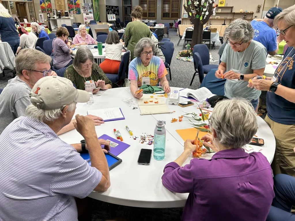 Retreat guests assemble orange flowers around a round table scattered with sequins and glue pens