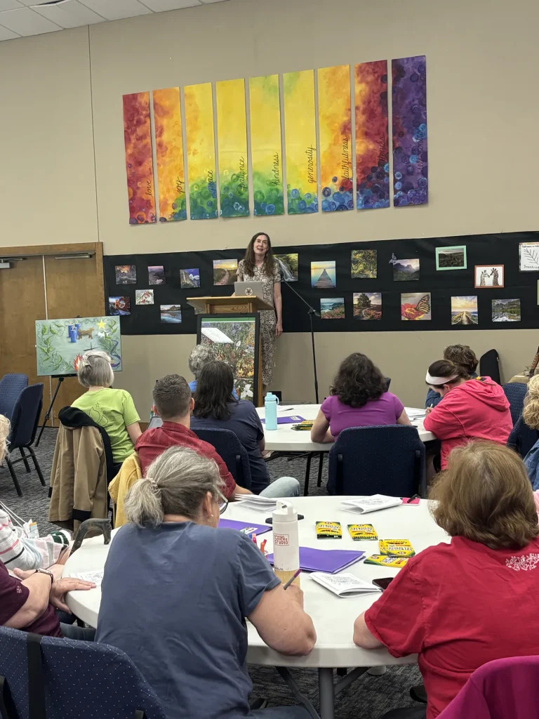Guests at round tables with crayons and coloring pages listen to a speaker beneath banners naming the fruits of the Spirit