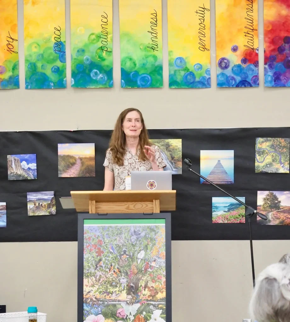 A speaker at a wooden podium beneath painted banners reading joy, peace, patience, kindness, generosity and faithfulness