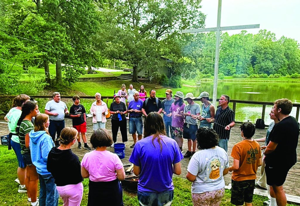 A service group gathered in a circle by the lake at Lutheridge, the cross beside the water