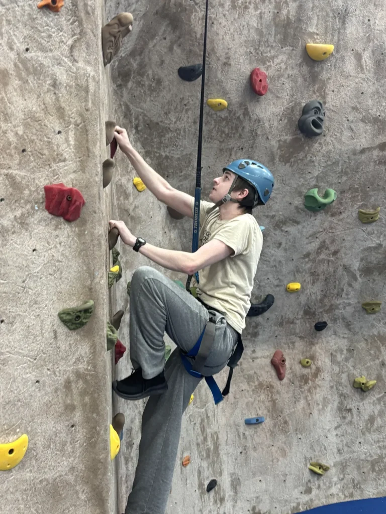 A teen in a blue helmet and harness climbing an indoor climbing wall