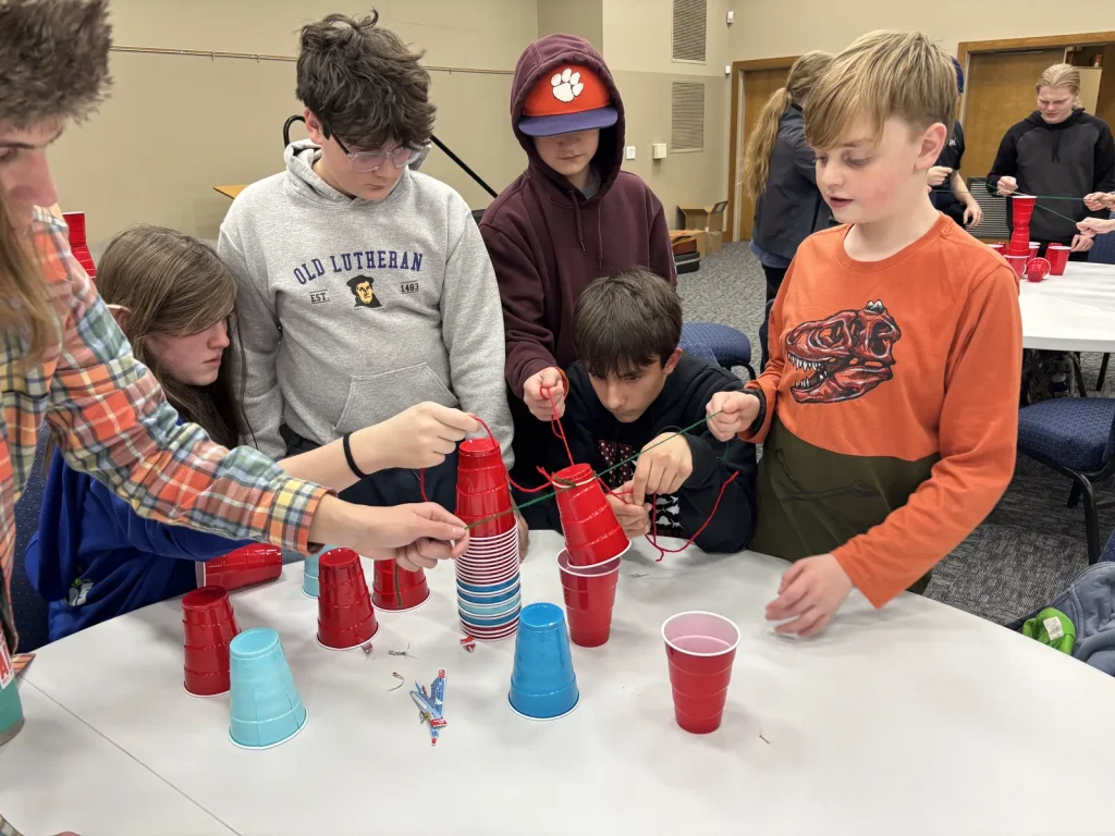 Youth around a table working together to stack red and blue cups with strings tied to a rubber band