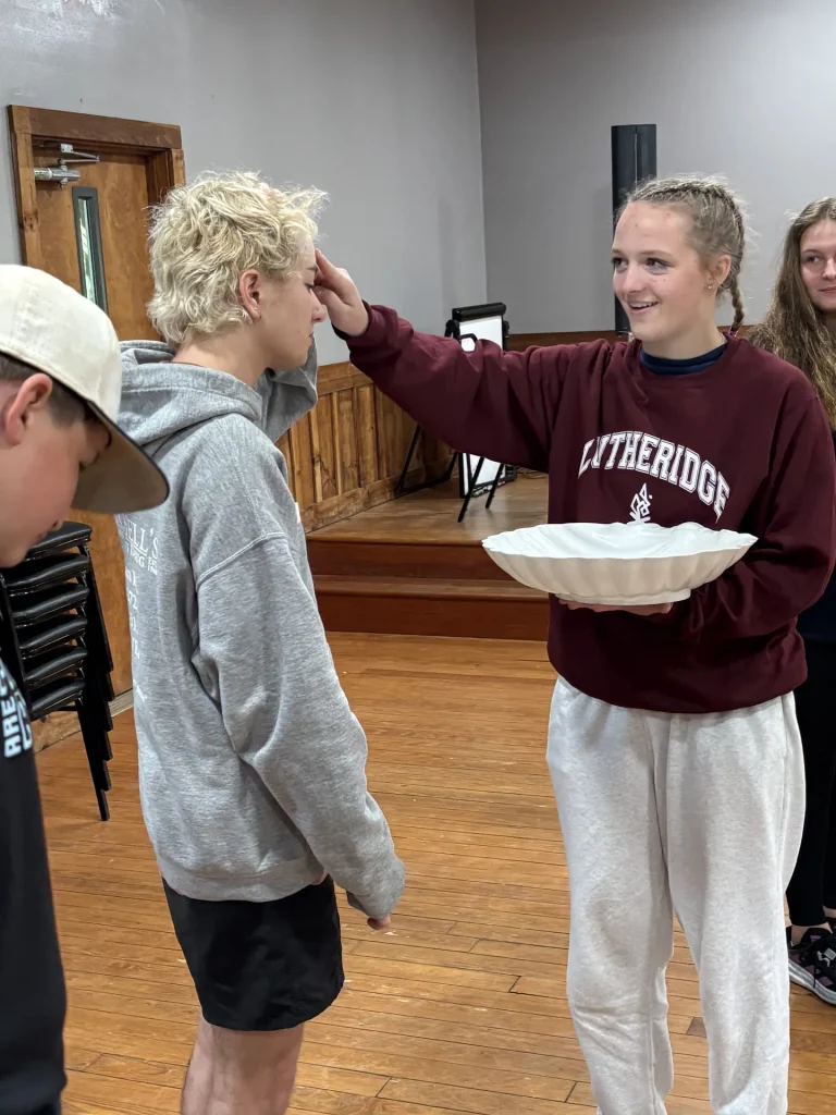 A counselor in a Lutheridge sweatshirt holds a shallow bowl and touches a camper's forehead