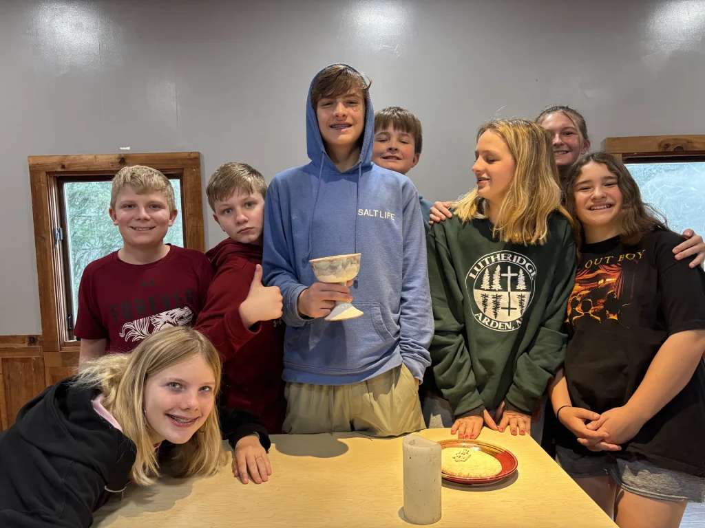 Eight smiling youth crowd around a table set with a chalice, a candle and a plate of bread, one in a Lutheridge sweatshirt