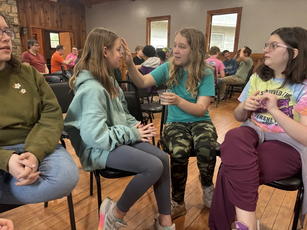In a circle of chairs, a girl reaches over to touch her friend's forehead