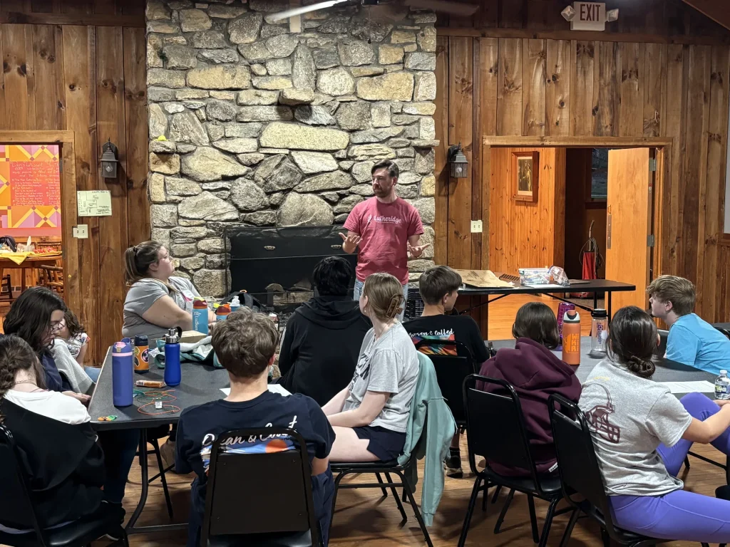 A leader in a Lutheridge T-shirt talks with youth at tables in front of a big stone fireplace in a wood-paneled lodge