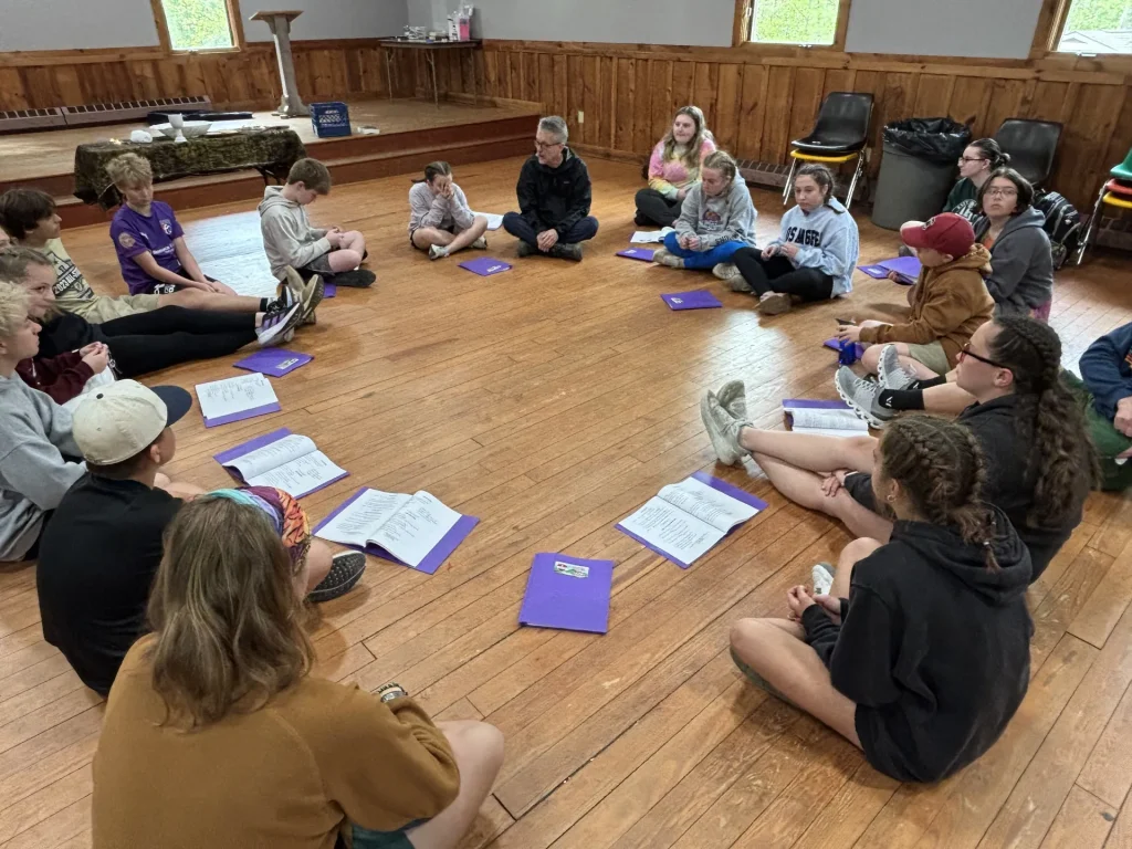 Youth and a leader sit in a wide circle on a wooden floor, purple folders open in front of them