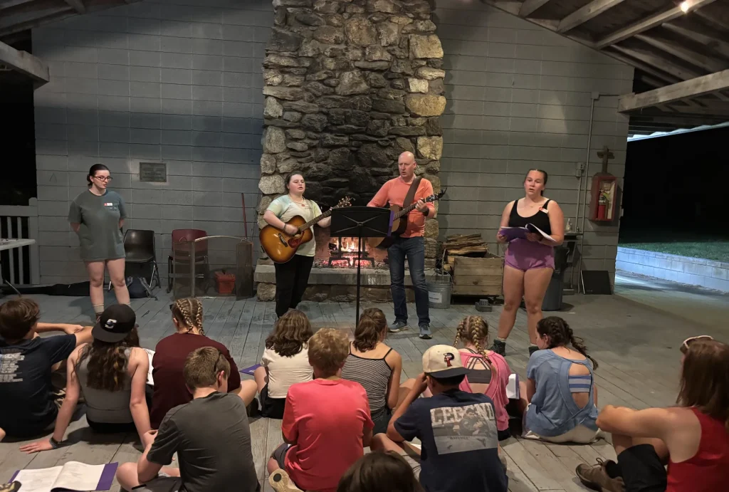 Youth sit on the floor of an open-air pavilion as two leaders with guitars lead an evening song beside a fire in the stone fireplace
