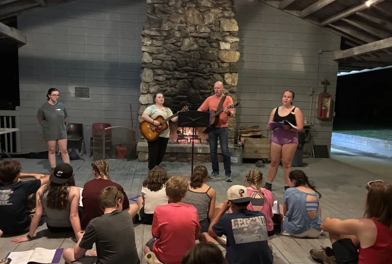 Youth sit on the floor of an open-air pavilion as two leaders with guitars lead an evening song beside a fire in the stone fireplace