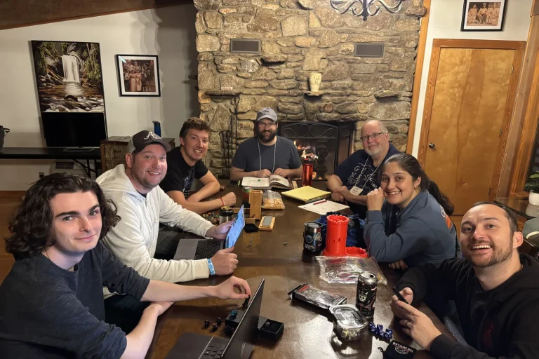 Geek Gathering guests smiling around a long game table by a stone fireplace, with dice, a dice tower and rule books spread out
