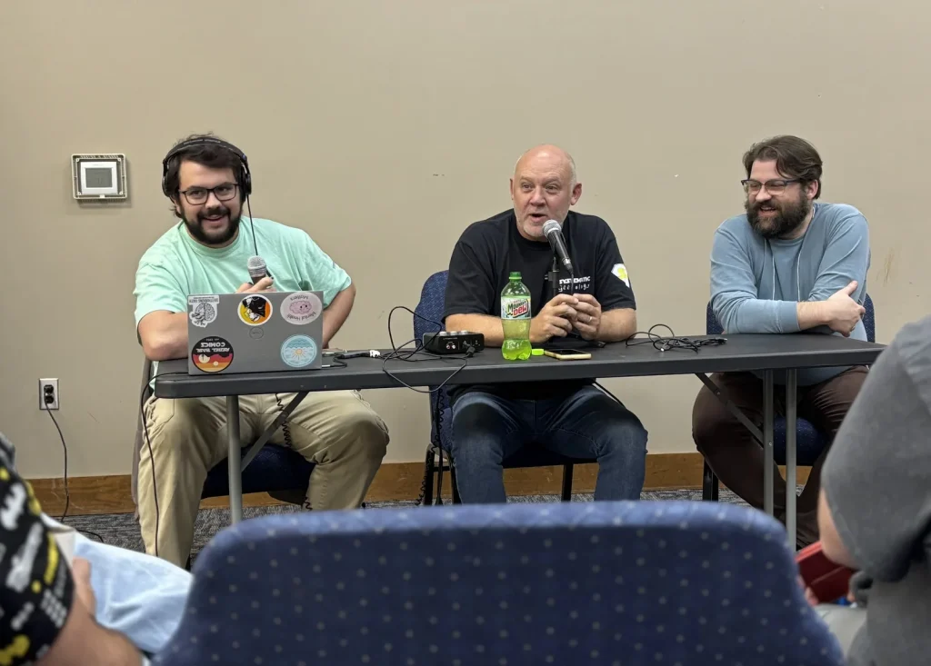 Three hosts at a table with microphones and headphones recording a live podcast