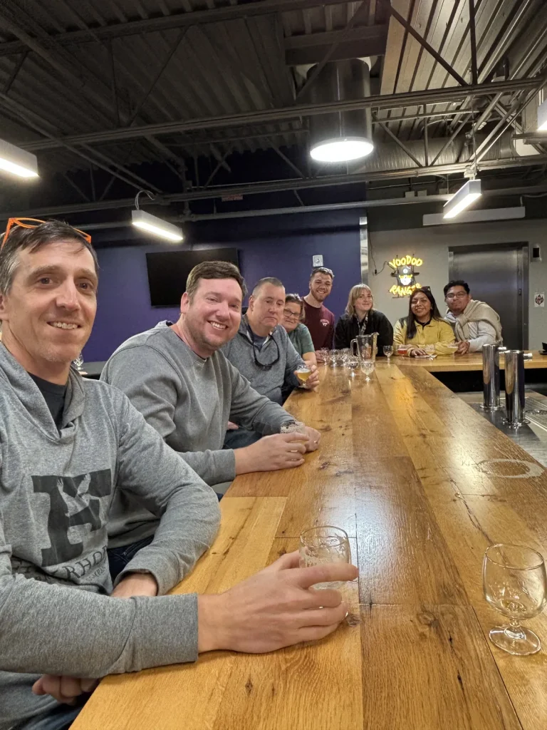 Theology On Tap guests smiling along a long wooden tasting table at a brewery