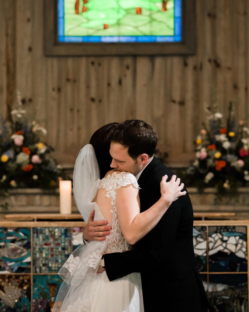 A bride and groom embrace at the altar, with flowers and stained glass behind them