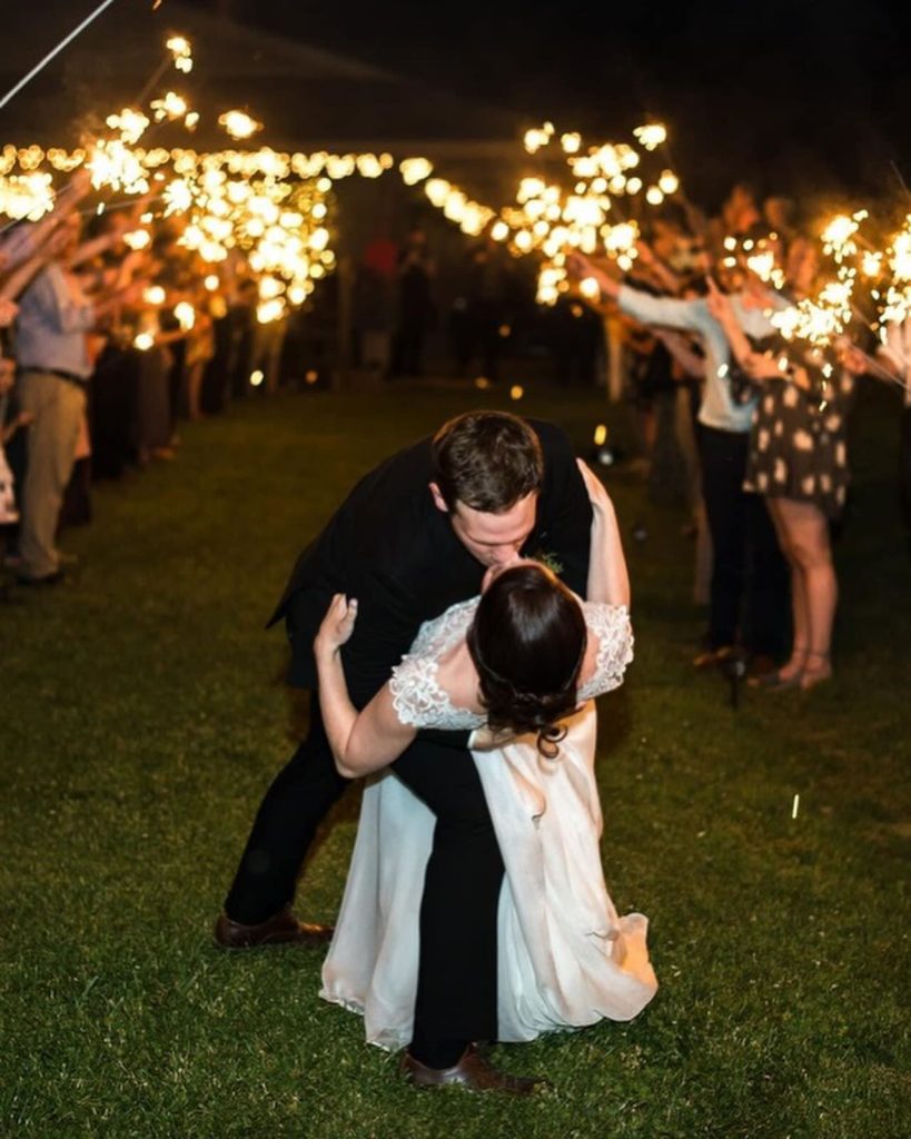 Guests hold sparklers high as the groom dips the bride for a kiss on the lawn at night