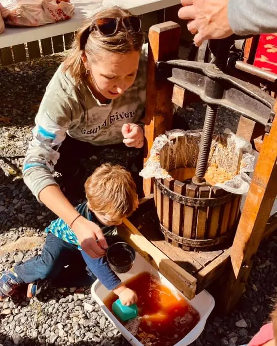 A young child and a woman work an old wooden cider press, fresh cider running into a tub