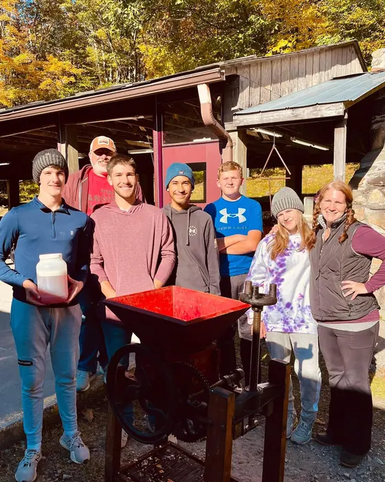 A family group stands by an apple grinder and cider press under a wooden shelter in the fall