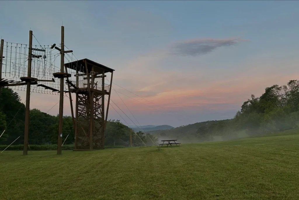 A ropes course tower at dusk over a misty field, mountains behind.