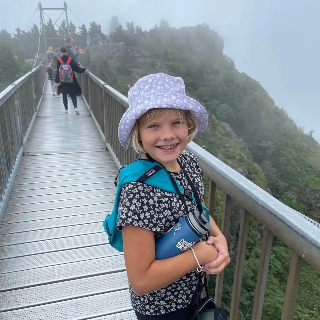 A smiling girl in a sun hat on Grandfather Mountain's swinging bridge, mist over the peaks behind her.