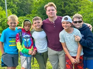 Five elementary-age campers and their counselor with arms around each other, smiling outdoors at Lutherock