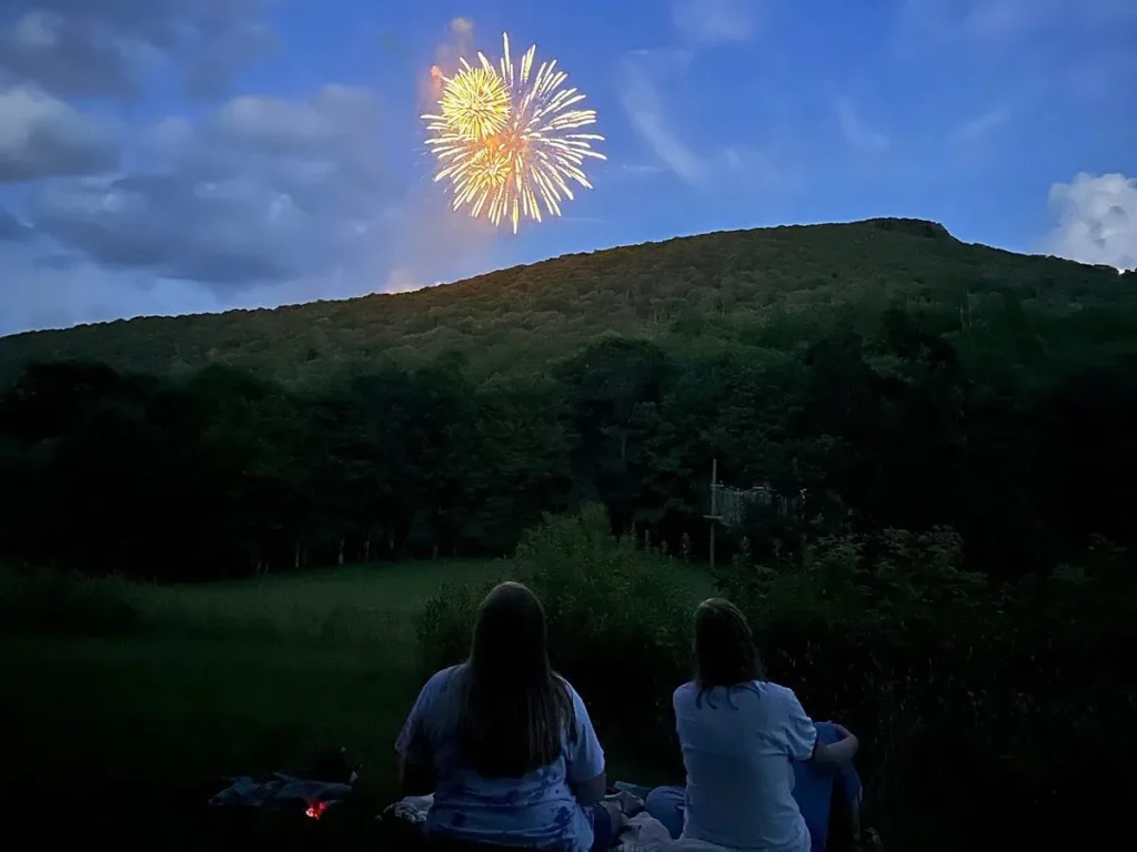 Fireworks bursting over the mountain at dusk while two people watch from a blanket on the Lutherock field