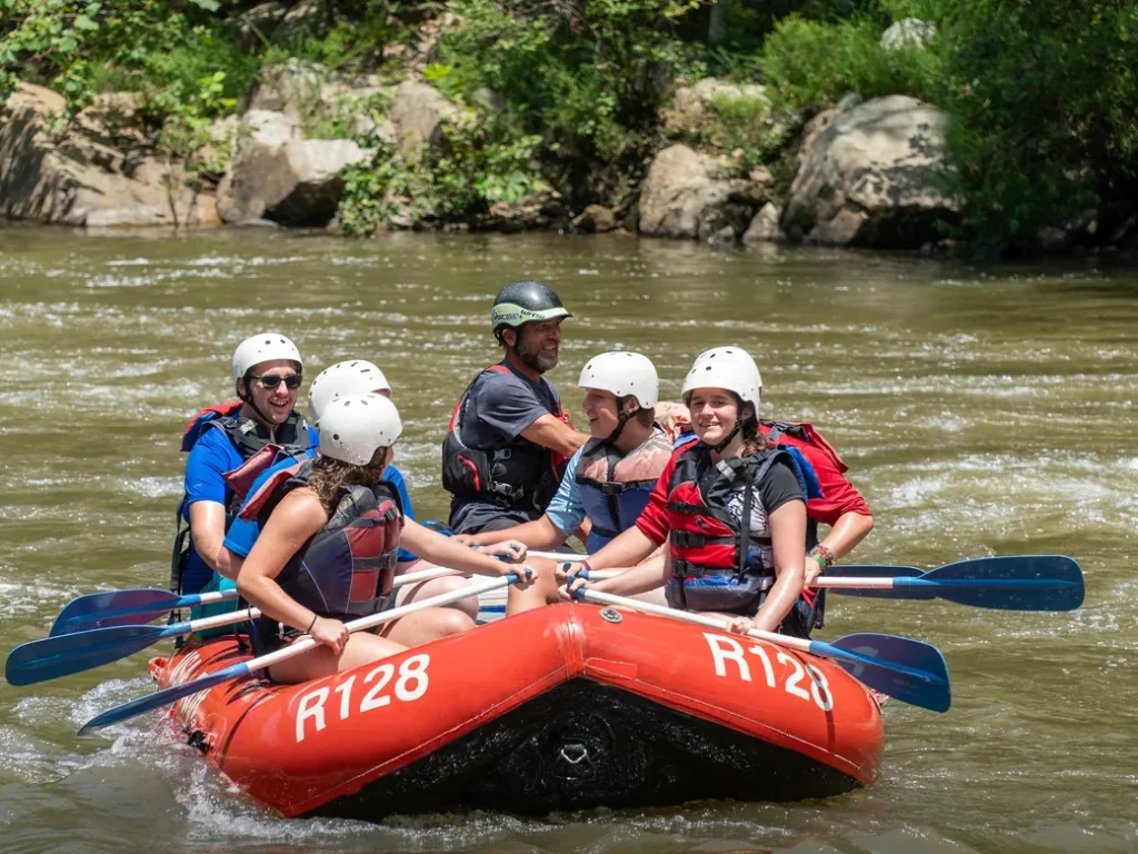 Campers in white helmets paddling a red raft through river rapids with their guide