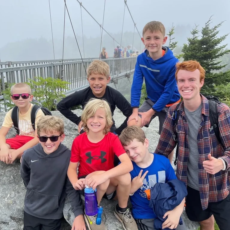 A smiling Lutherock counselor giving a thumbs up with a group of young campers on a mountain outing, a suspension bridge behind them