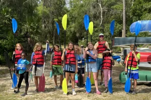 A cabin group of young campers in life jackets holding their paddles up beside the canoe racks at Luther Springs