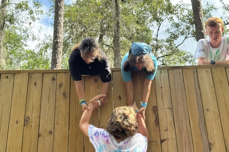 Two teens lean over the top of a tall wooden wall to pull a teammate up by the hands, a team challenge in the pines at Luther Springs
