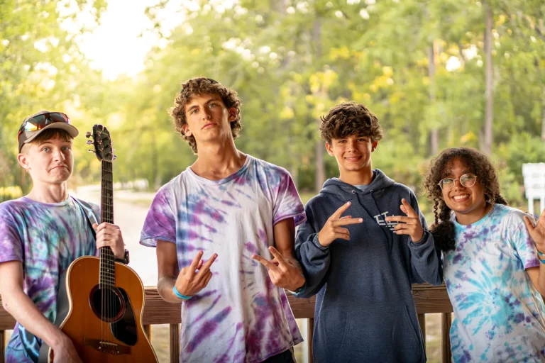 Four Leadership Lab teens in tie-dye shirts, one holding a guitar, grinning outdoors at Luther Springs