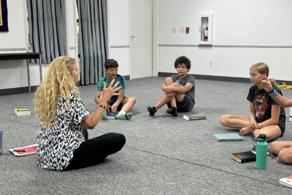 A leader sits on the floor with a circle of middle schoolers and their Bibles, talking with her hands