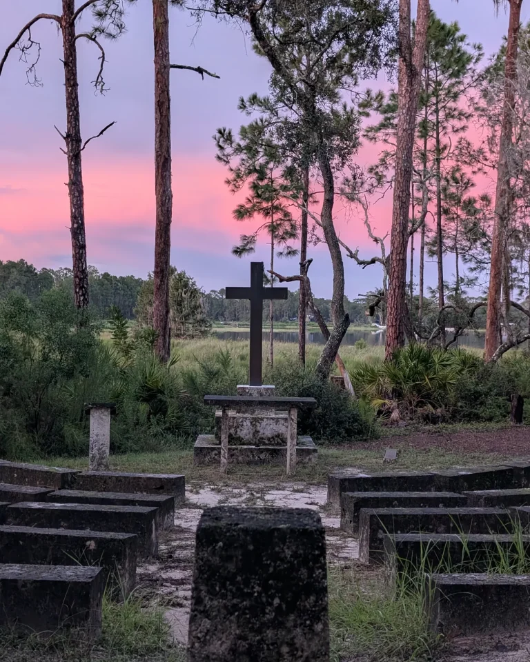 A wooden cross at the front of the outdoor chapel at Luther Springs, framed by pines and palmettos under a pink sunset sky